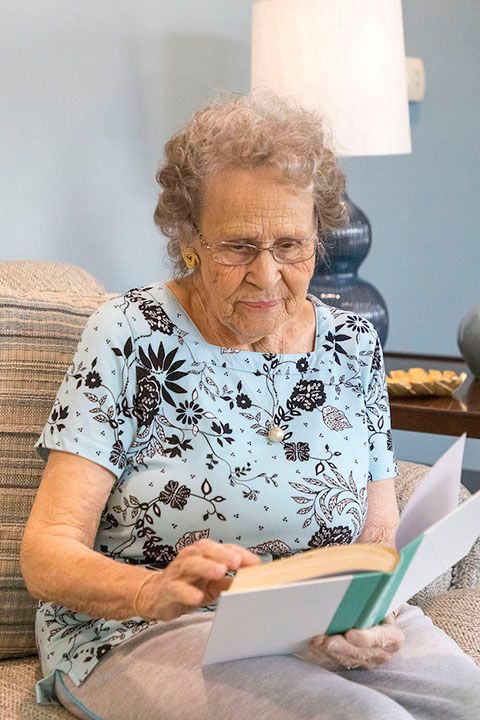 An elderly woman is sitting on a couch reading a book.