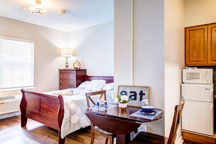 A bedroom with a bed , table , chairs and a refrigerator.