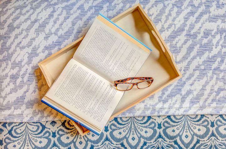 A book and glasses are on a wooden tray on a bed.
