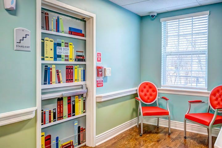 A waiting room with two red chairs and a bookshelf filled with books.