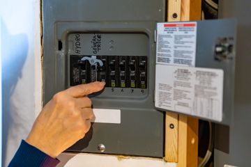 Person's hand pointing at circuit breaker in electrical panel, gray box in wooden frame.
