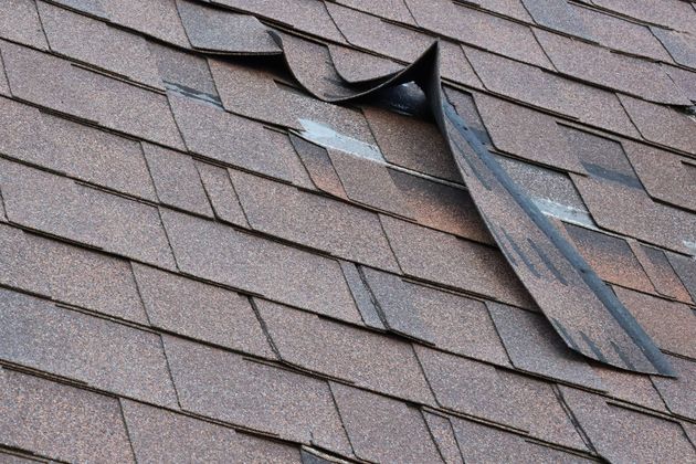 Damaged brown asphalt roof shingles with a section torn and curled up.