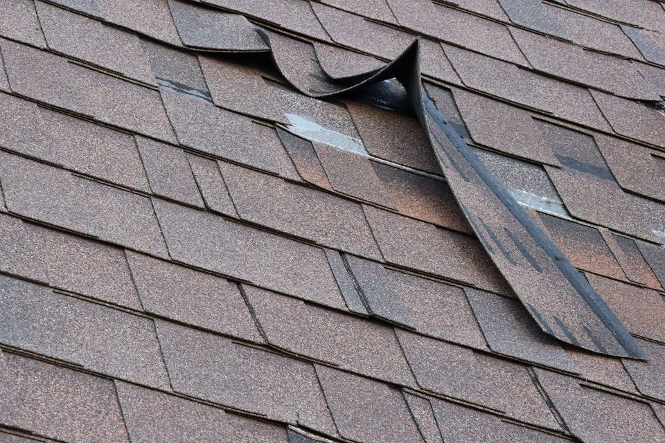 Damaged brown asphalt roof shingles with a section torn and curled up.