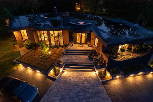 Night view of a modern brick home with illuminated walkway and landscaping. A car is parked in the driveway.
