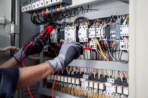 Electrician testing electrical panel with a multimeter. Black gloves, wires, and equipment visible.
