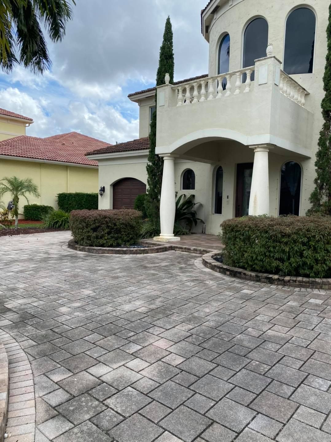 Brick-paved driveway leads to a large, light-colored stucco house with a balcony and arched windows under a cloudy sky.