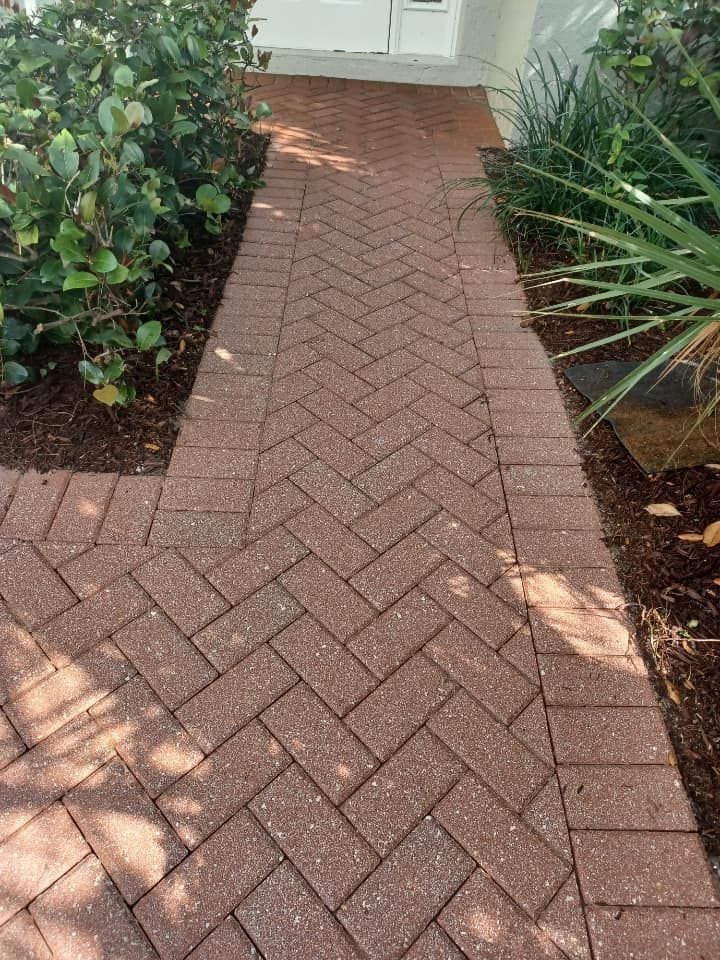 Red brick walkway in a herringbone pattern, leading to a white door. Lush green bushes border the path.