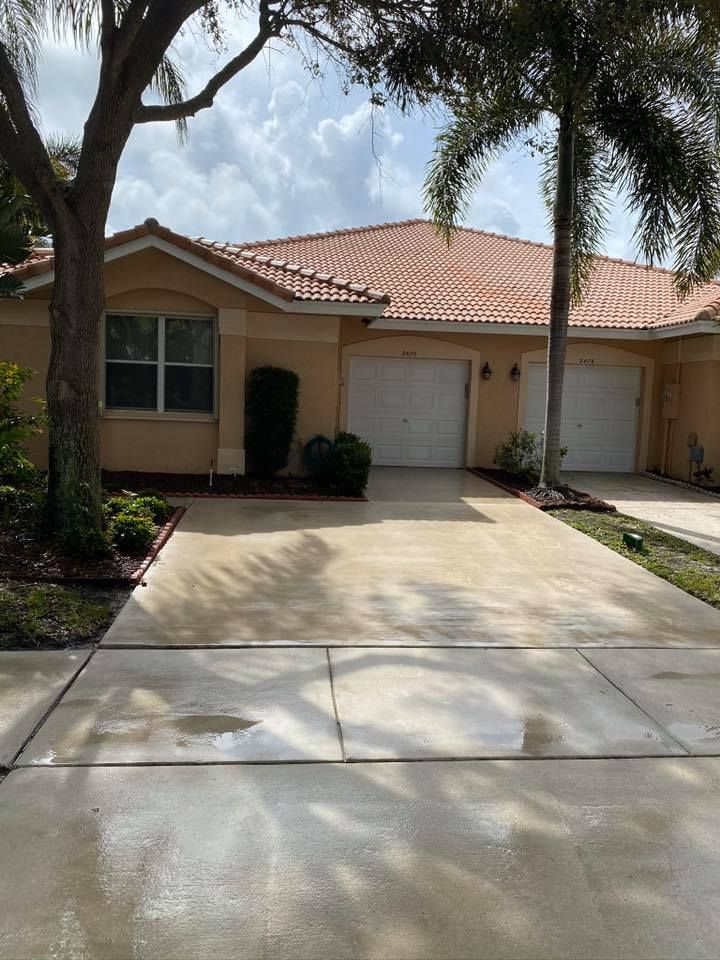 Tan townhouse with red tile roof and two garage doors. Driveway in front with trees and cloudy sky overhead.