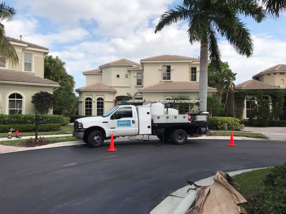 A white service truck parked on newly paved asphalt in front of large houses. Orange traffic cones mark the work area.