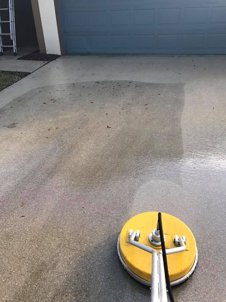 Pressure washer cleaning a concrete driveway. The circular yellow cleaner is removing dirt, leaving lighter areas.
