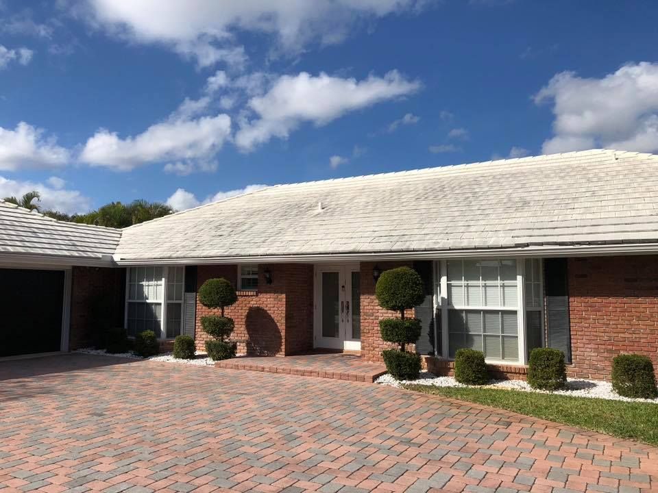 A single-story brick house with a white roof, surrounded by a brick driveway and green bushes under a partly cloudy sky.