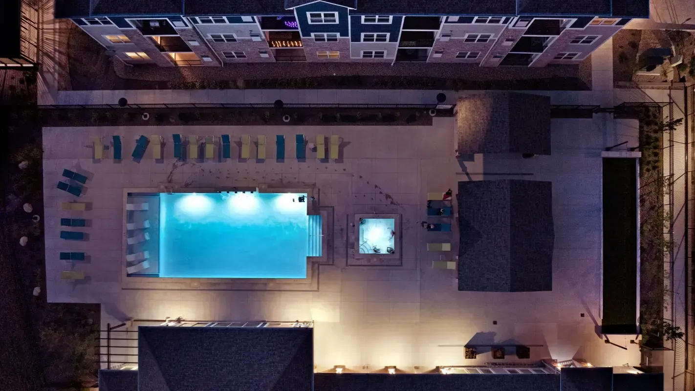 Aerial night view of an apartment community pool with lounge chairs and surrounding buildings.
