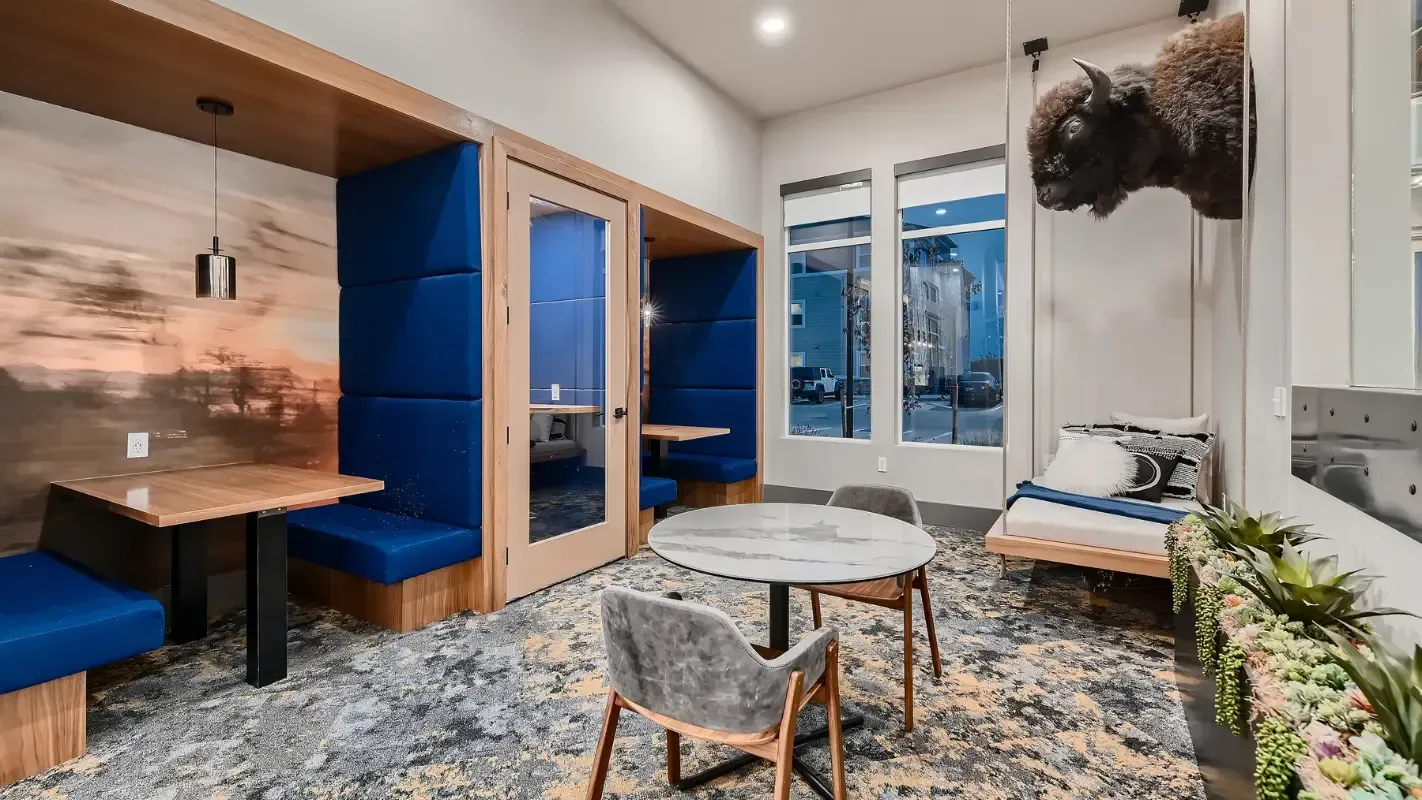 Interior of a community lounge: blue padded booths, round marble table, and large windows.