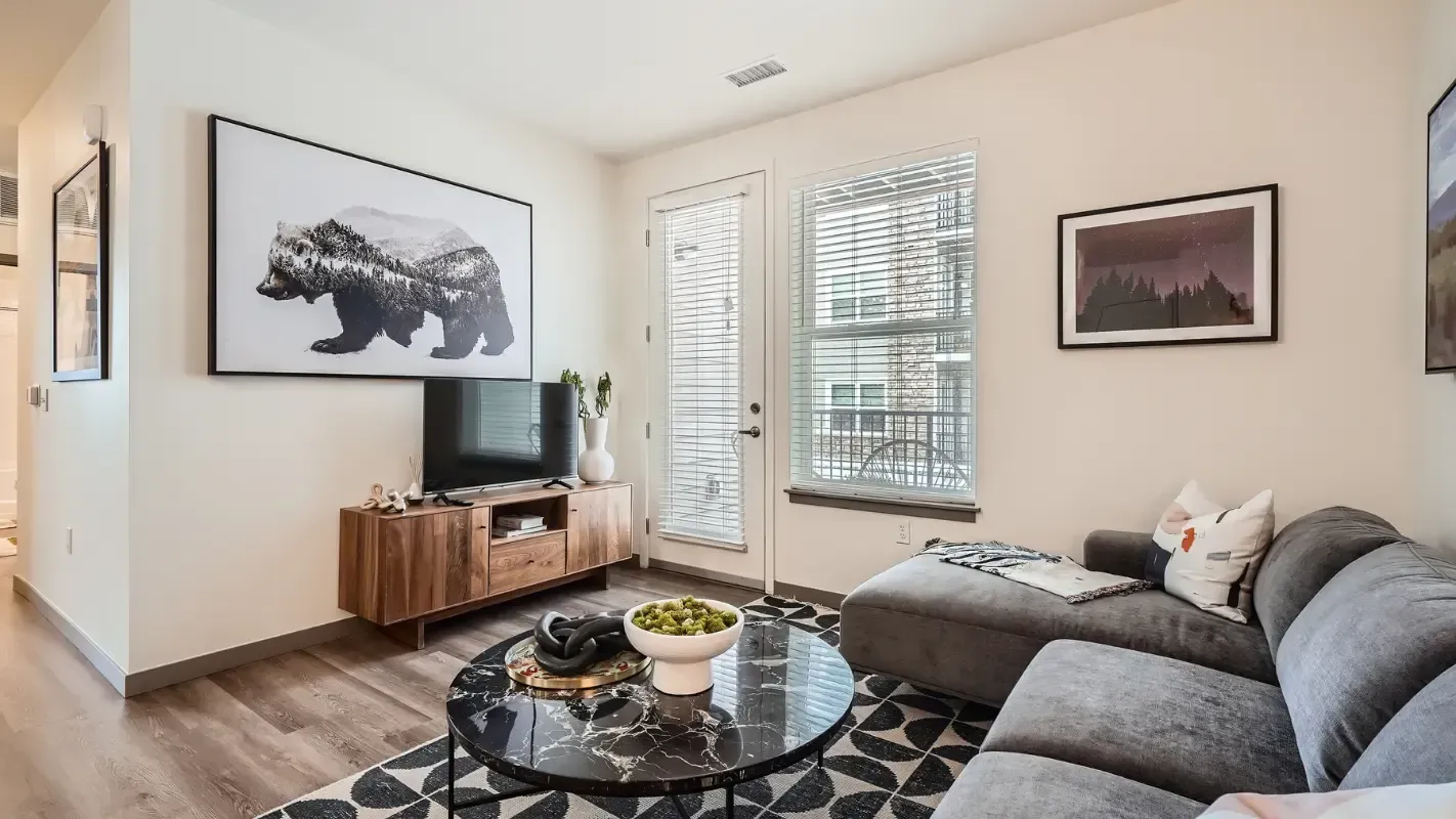 Living room in a modern apartment with a gray sectional, round marble coffee table, TV console, and large windows.