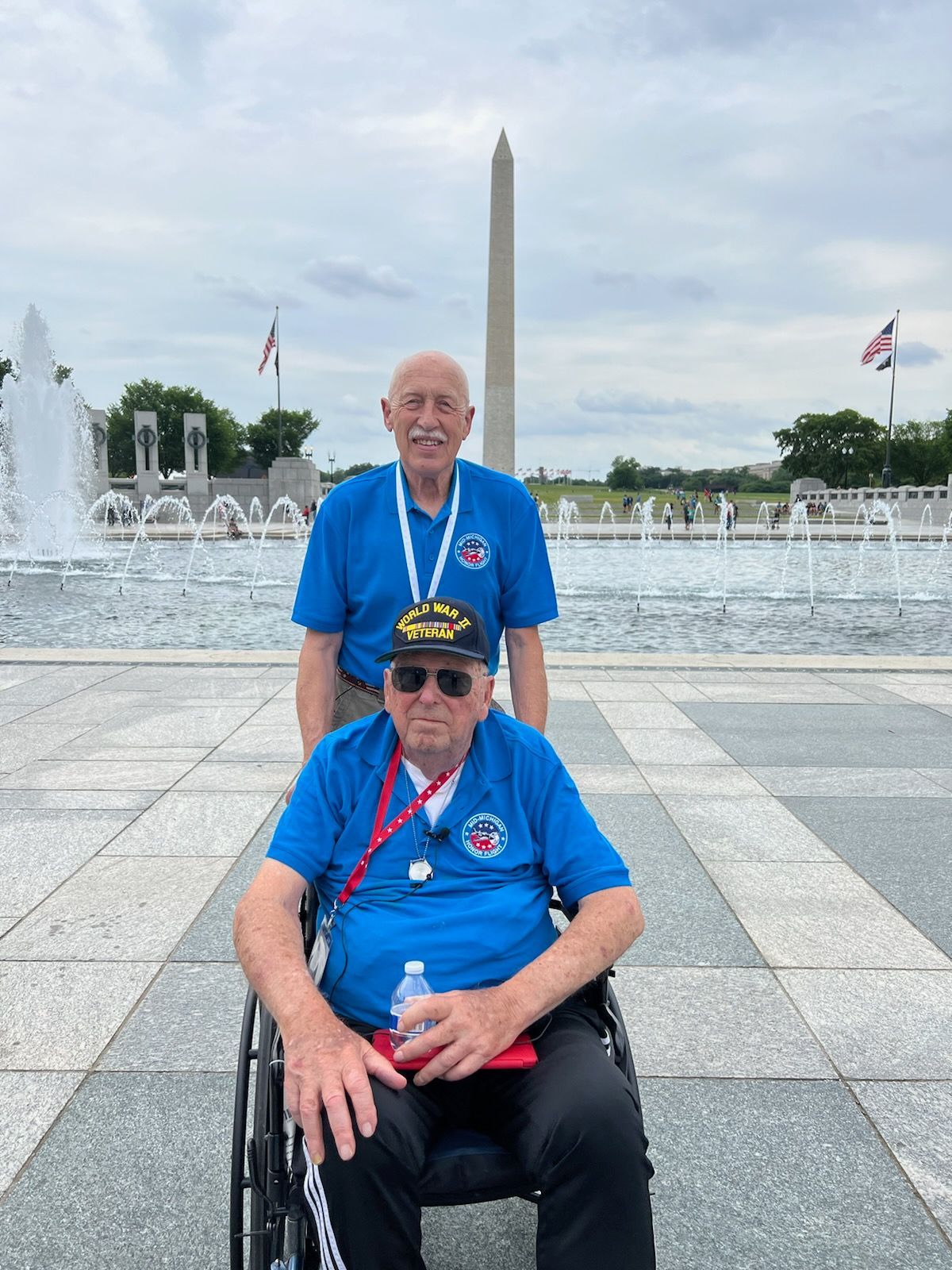 A man in a wheelchair is standing next to another man in front of a fountain.