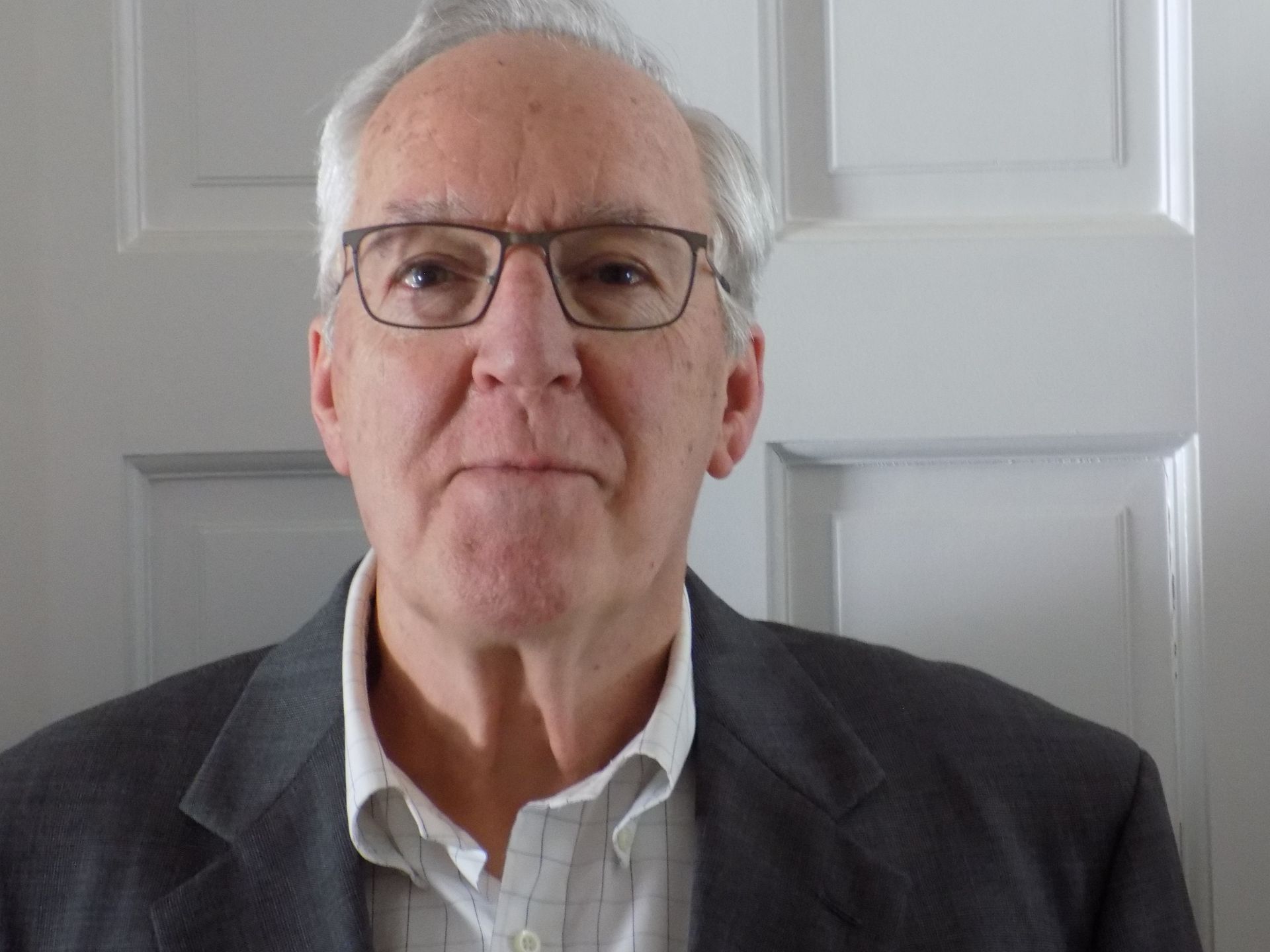A man wearing glasses and a suit is standing in front of a door.