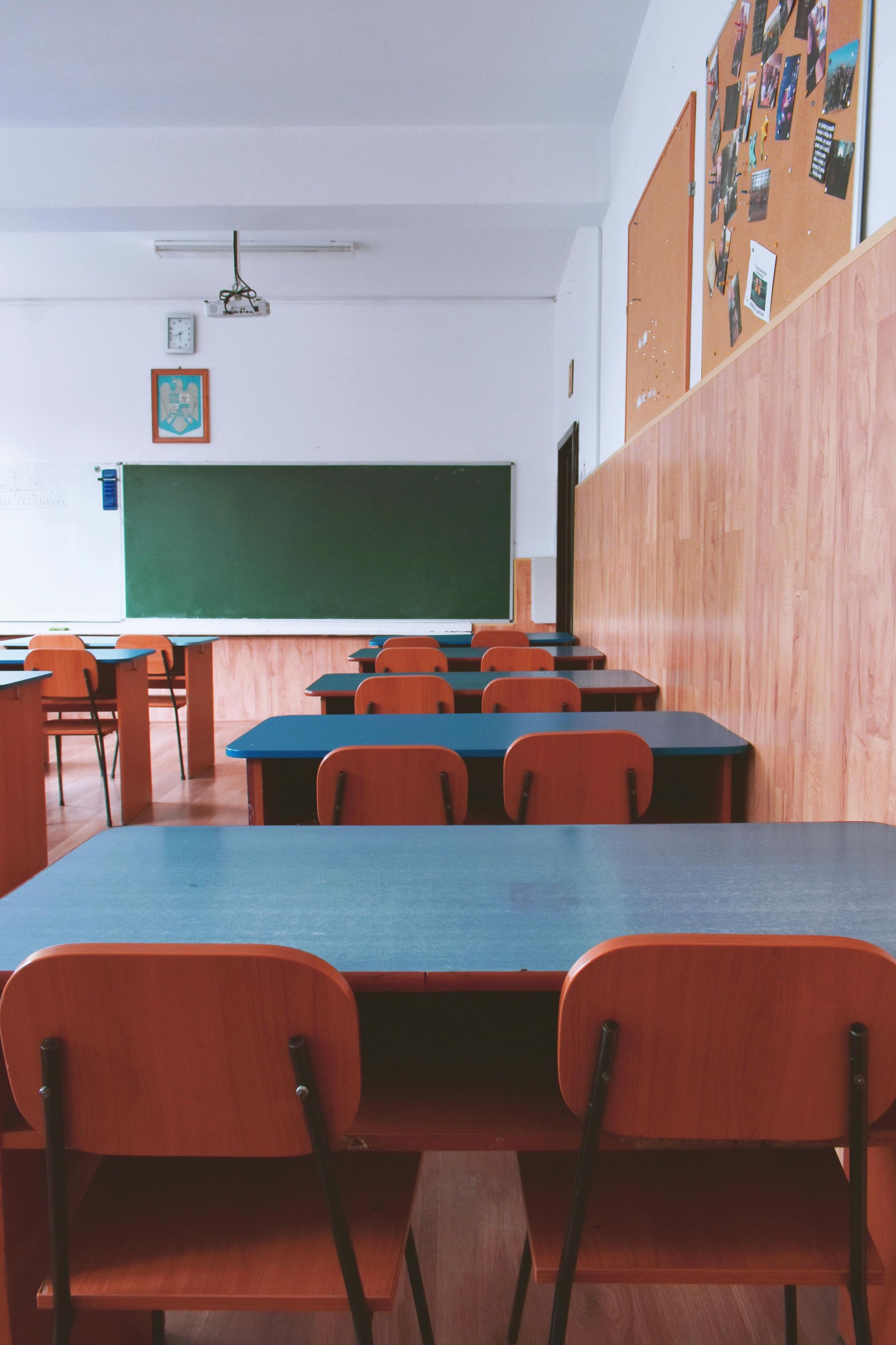 Empty classroom with rows of desks and chairs facing a chalkboard. Wooden paneling lines the walls.