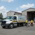 Recycling truck with workers loading materials near a sorting facility on a sunny day.
