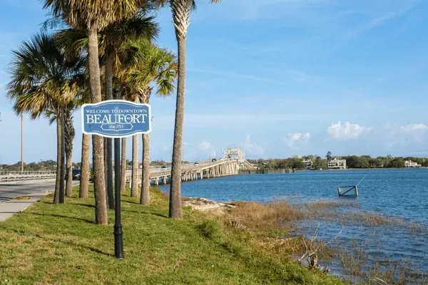 Beaufort, SC welcome sign with waterfront view, bridge, and palm trees under a blue sky.
