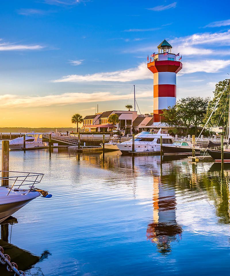 Red and white striped lighthouse at harbor, reflecting in calm water, boats docked, blue sky. Hilton Head Island