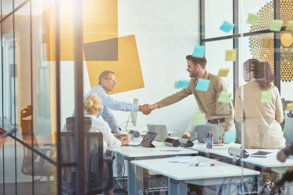 People shaking hands in an office meeting. Yellow and blue sticky notes on the glass wall.