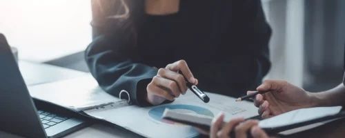 Two people reviewing documents with pens at a table, near a laptop.