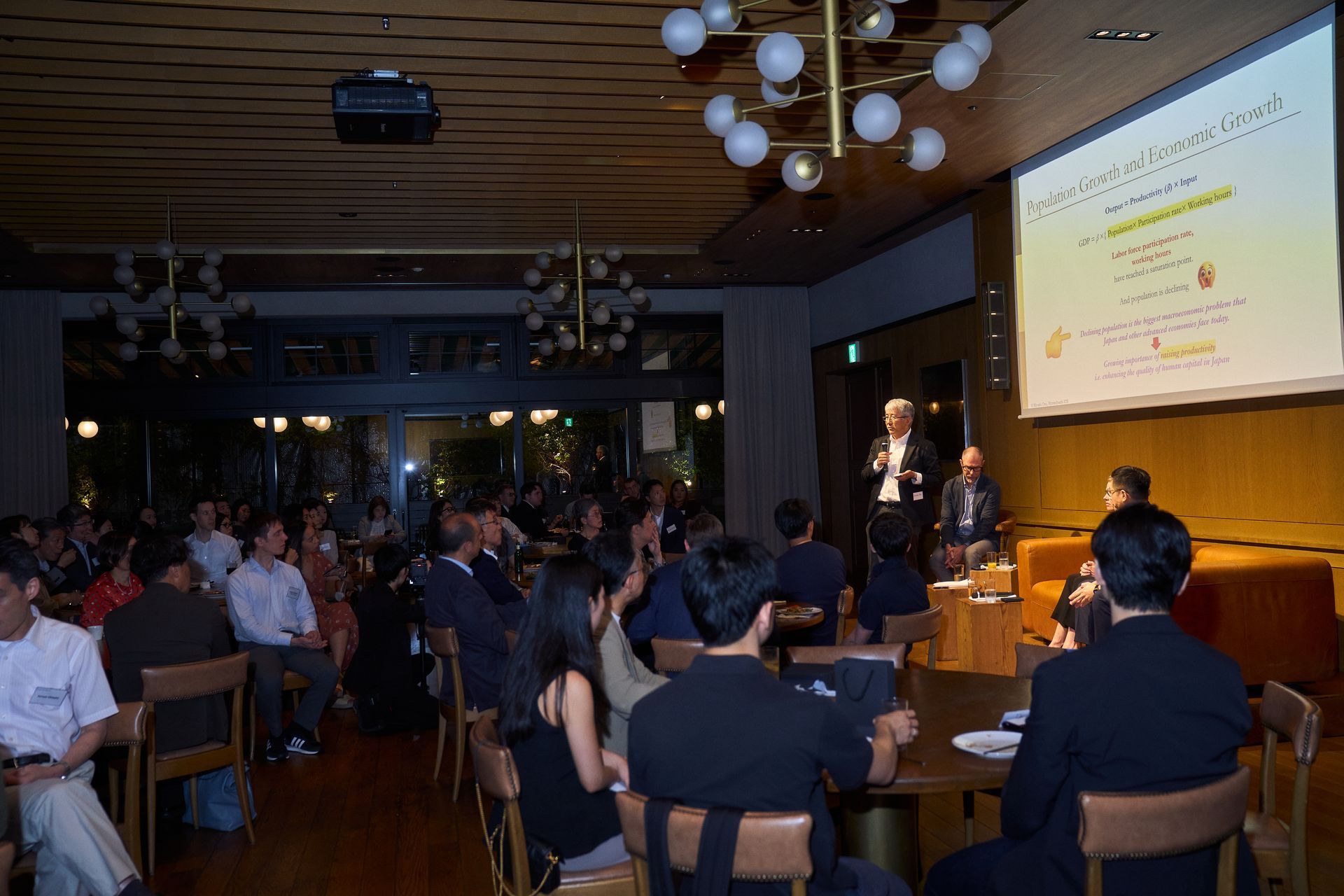 Audience watches a presentation in a dimly lit conference room with a projected slide on the wall