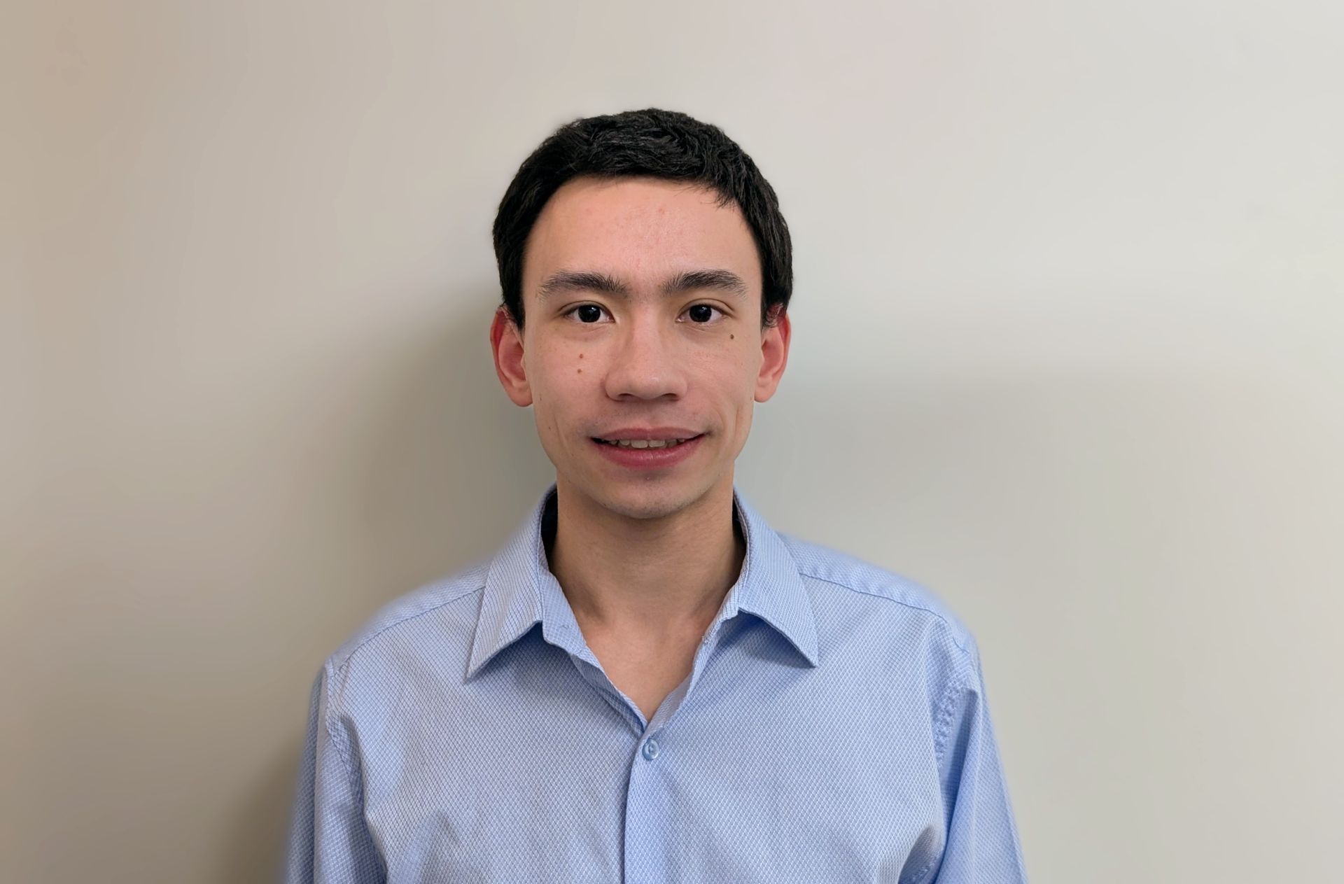 A young man in a blue shirt is standing in front of a white wall.