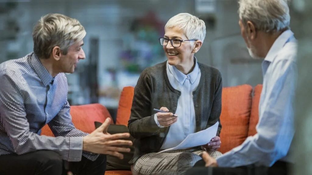 Three people in discussion on an orange couch, one smiling with documents, the others gesturing.