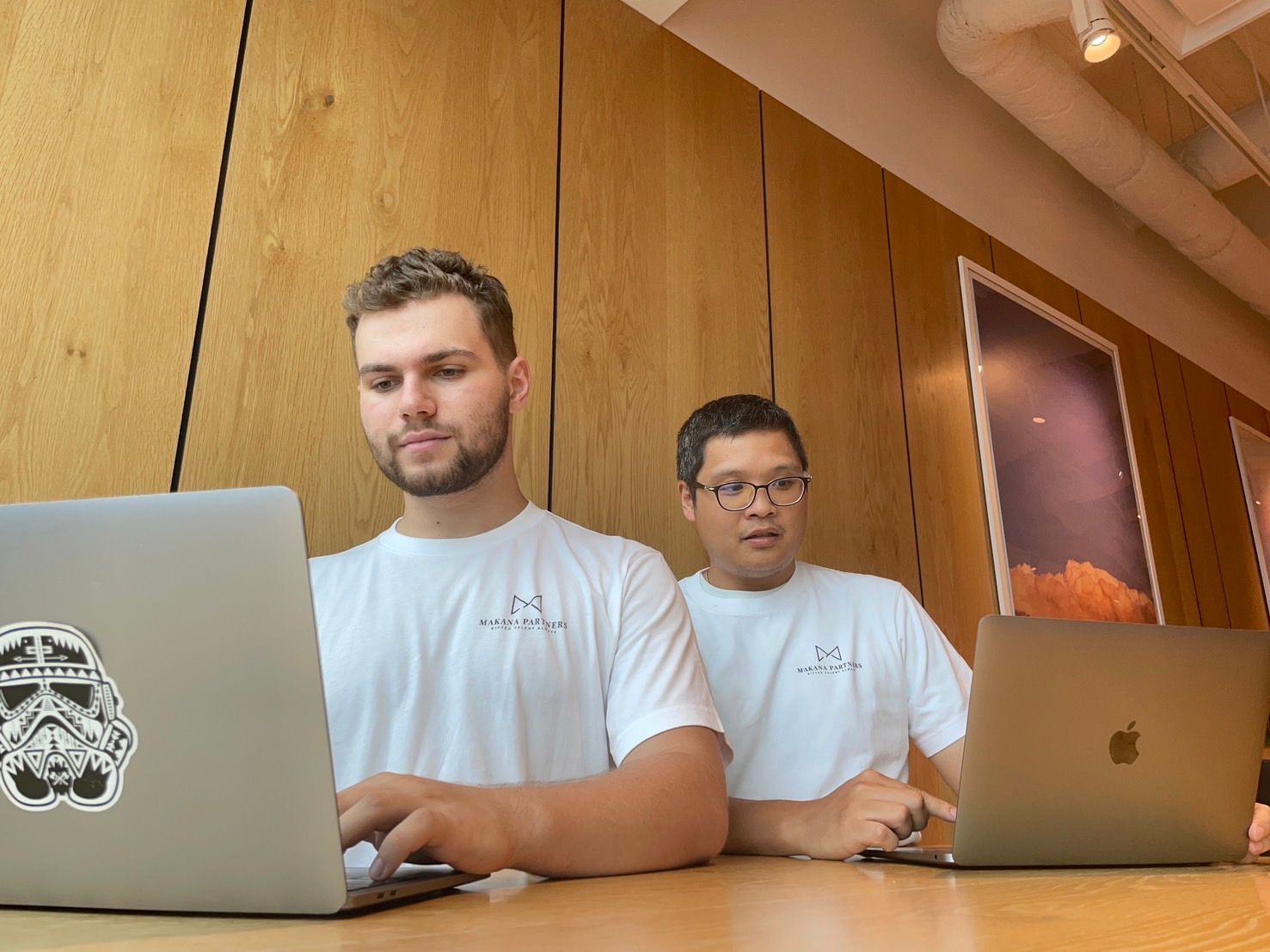 Two men are sitting at a table using laptops.