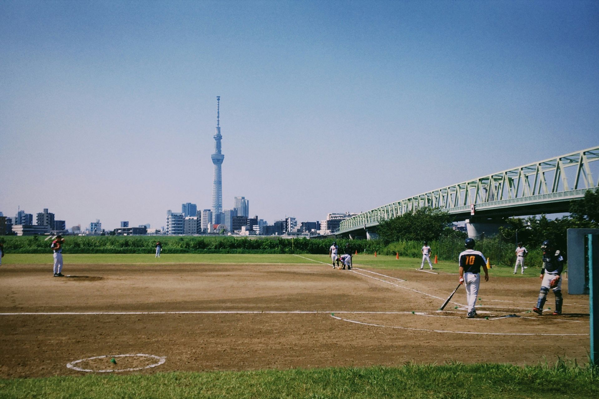 A group of people are playing baseball on a field with a bridge in the background.