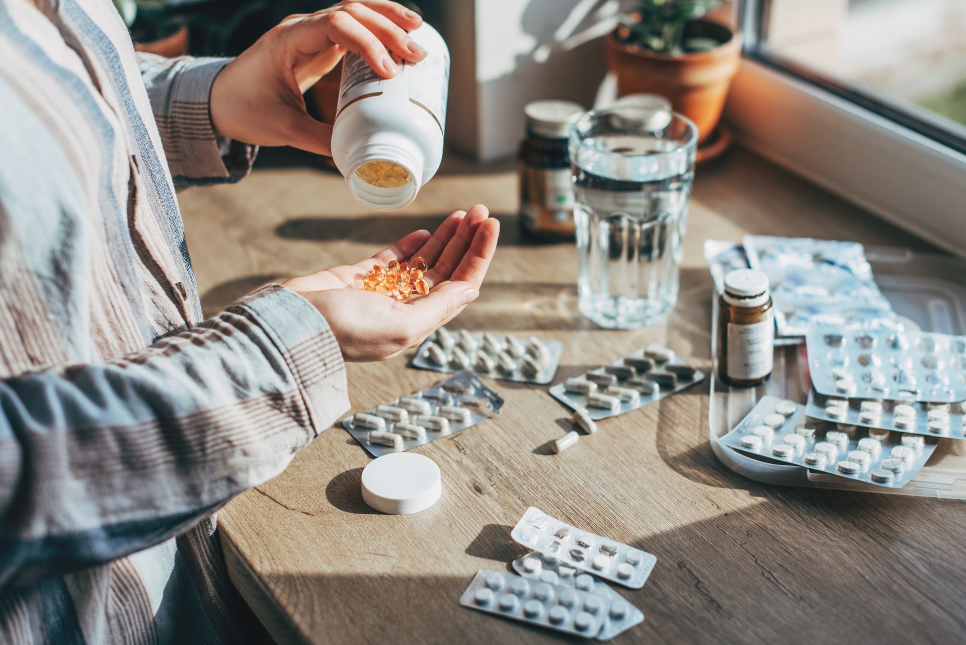 Person pouring pills from a bottle into their hand near medication and a glass of water on a wooden surface.