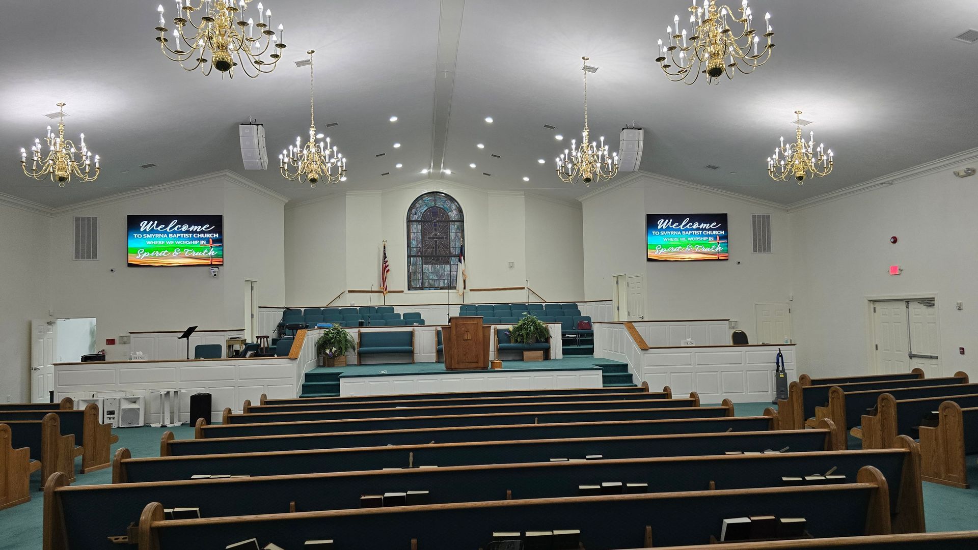 Interior of a church sanctuary with rows of pews facing a raised stage. Two screens display text, and chandeliers hang from the ceiling.