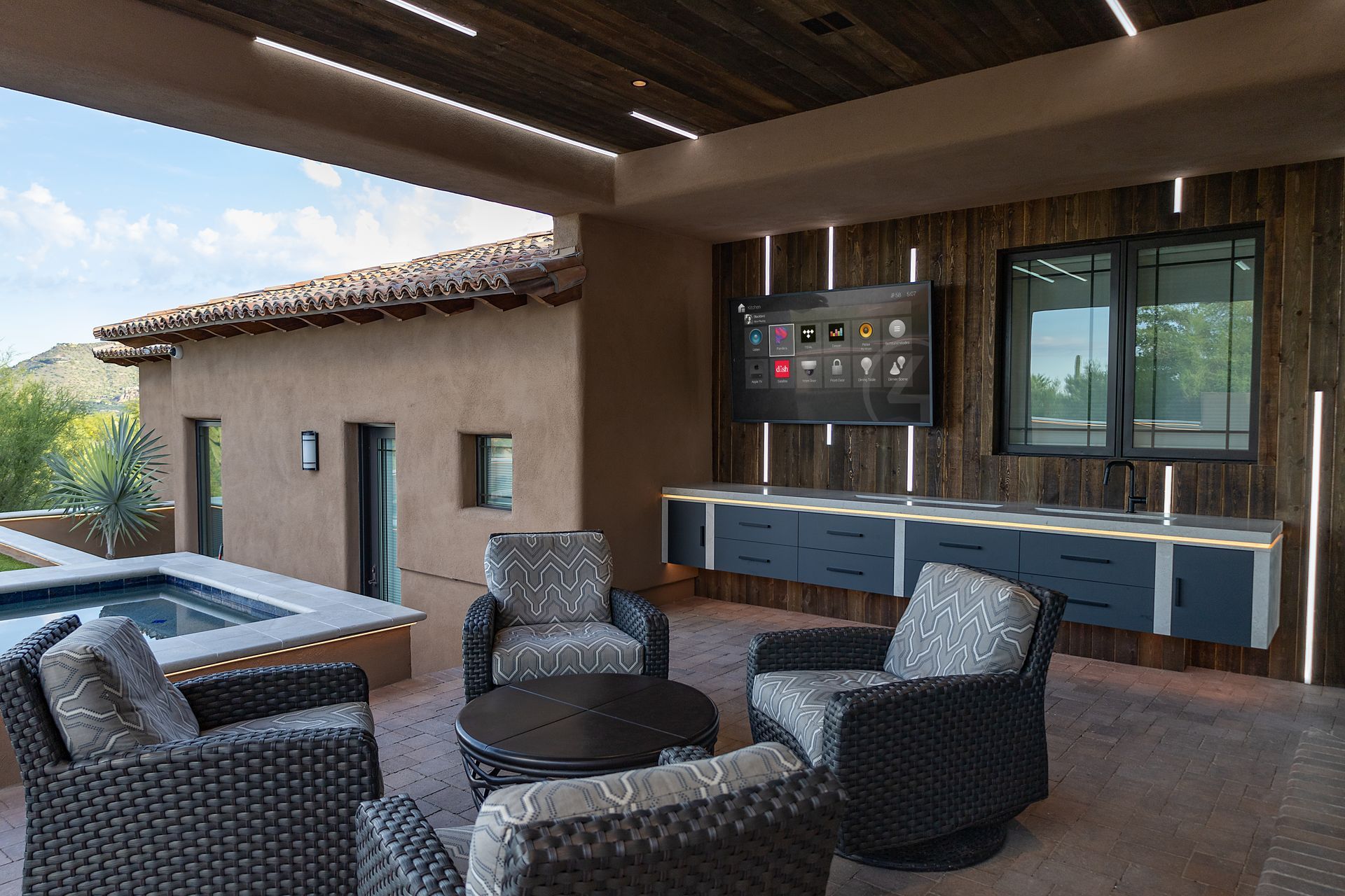 Patio with seating area and TV mounted on a wooden wall, overlooking a pool and distant view.
