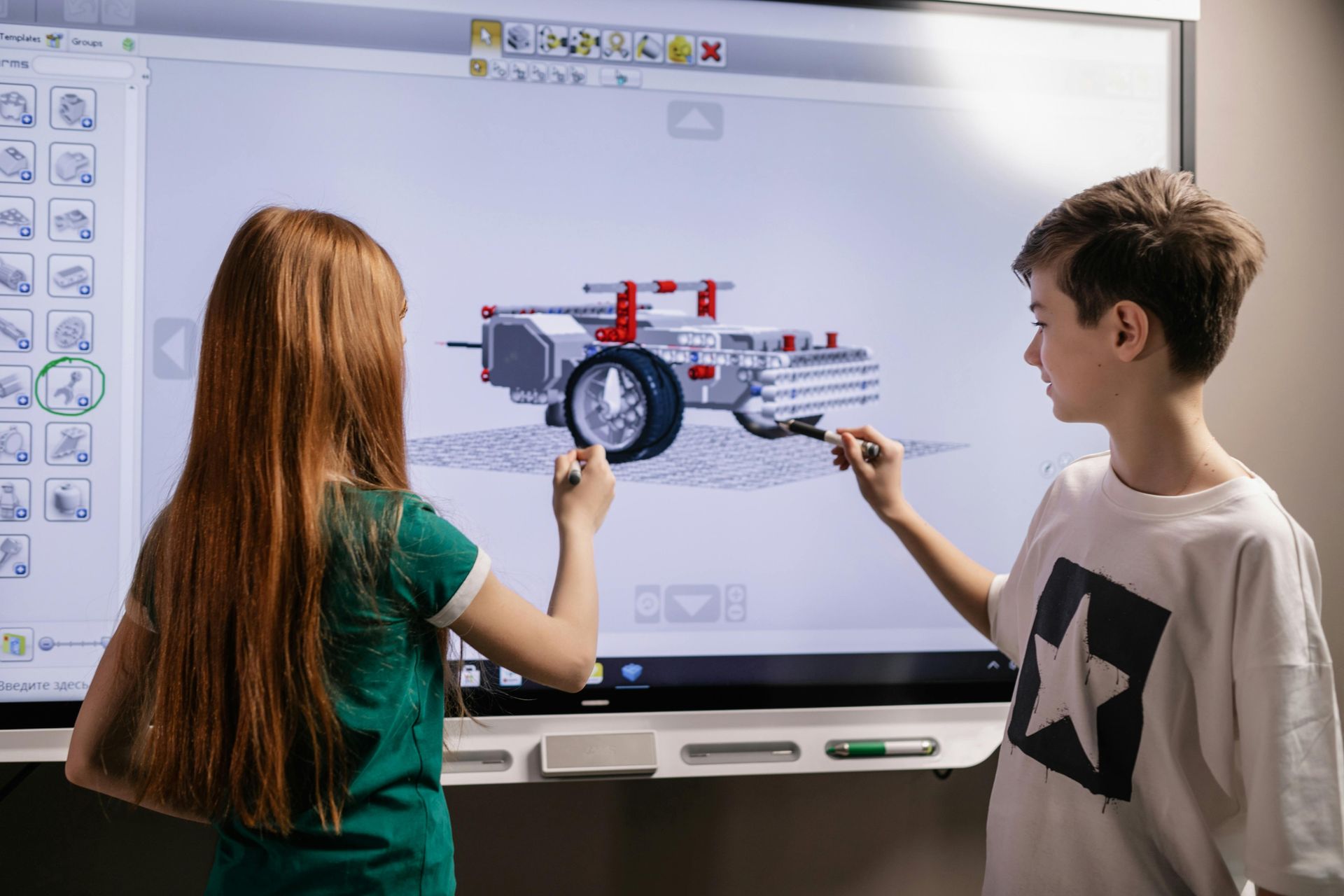 Two children use touch pens on a large screen showing a 3D model of a Lego vehicle.