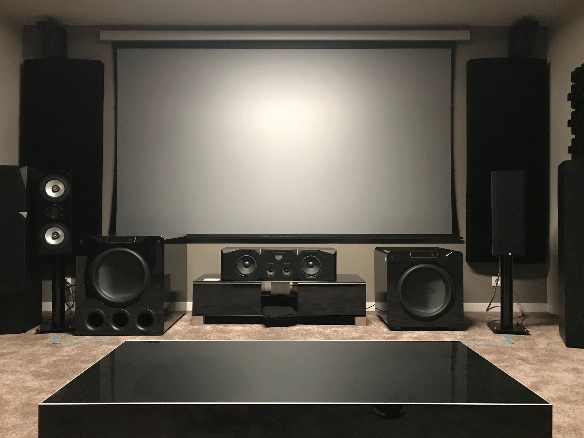 A home theater setup with a projector screen, speakers, and a black coffee table. The room is dark, and speakers are placed around the screen.