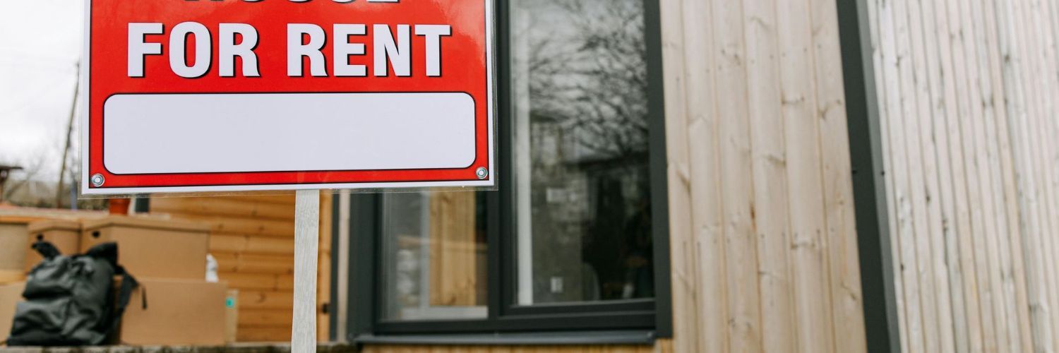 A red for rent sign is in front of a wooden house.