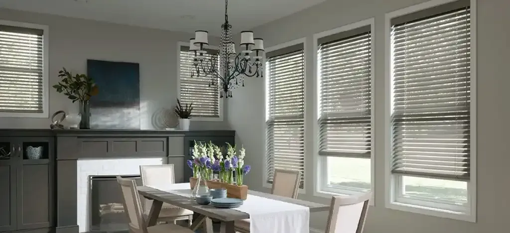 Dining room with table set for a meal; windows with blinds let light in. Gray, neutral tones.
