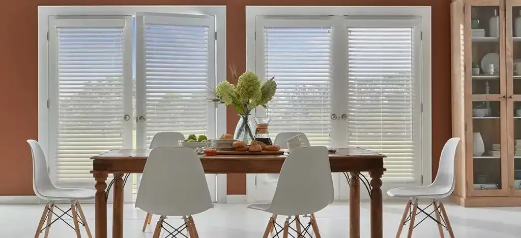Dining room with wooden table, white chairs, and glass-paned windows with blinds.
