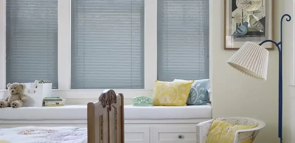 A cozy window seat with cushions, blinds, and a teddy bear. A floor lamp and artwork are nearby.