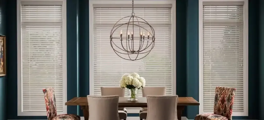 Dining room with a table, chairs, and a chandelier, framed by three large windows with blinds. Teal walls.