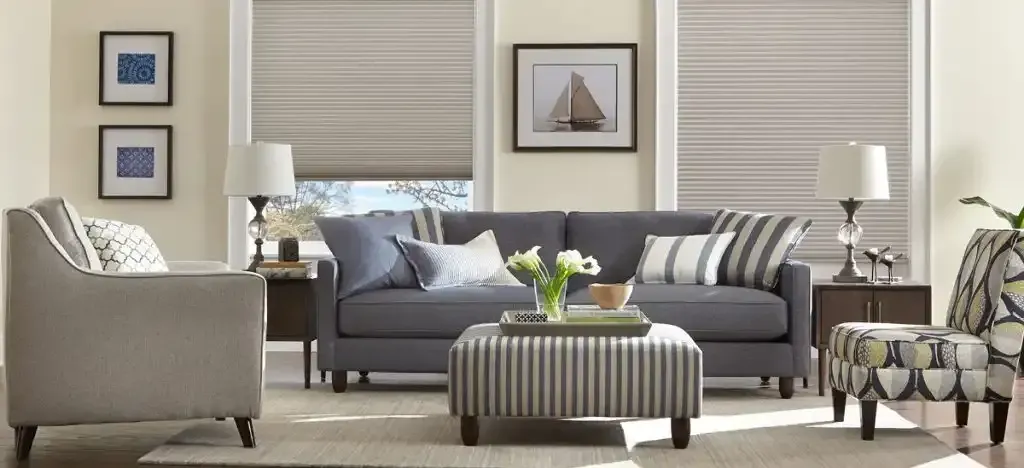 Living room with gray sofa, patterned chairs, striped ottoman, lamps, art, and neutral-toned window shades.