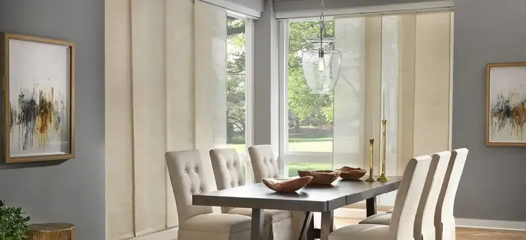 Dining room with table, chairs, art, and cream-colored panel shades. Grey wall.