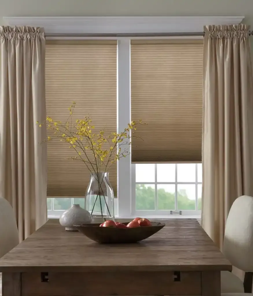 Dining room window with beige curtains, honeycomb blinds, wooden table, and vase of flowers.