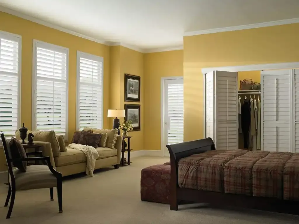 Bedroom with yellow walls, white shutters on windows, a bed with a brown plaid comforter, and a sofa.