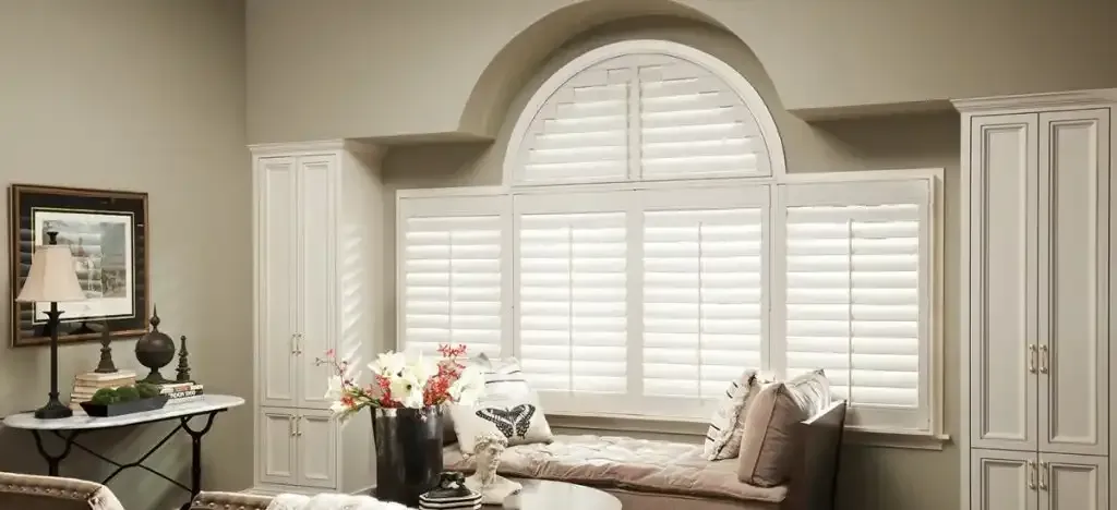 A sunlit room with white shutters on a large arched window, a built-in window seat, and neutral-toned walls.