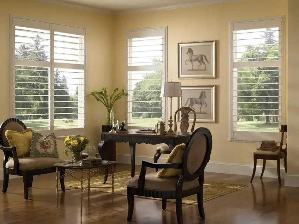 Living room with shutters, desk, chairs, and artwork; warm yellow walls and hardwood floors.