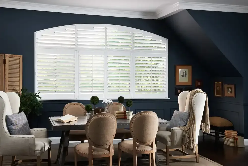 A dining room with white shutters on a large arched window. Navy blue walls, a table, and chairs are visible.