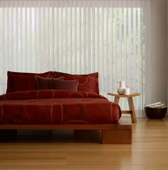 Bedroom with red bedding, wood platform bed, small table with vases, vertical blinds.