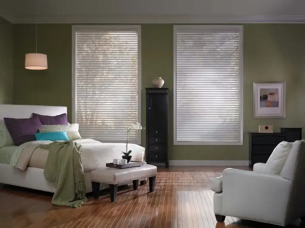 Bedroom with a bed, chair, and two windows with blinds. Olive green walls, hardwood floor.